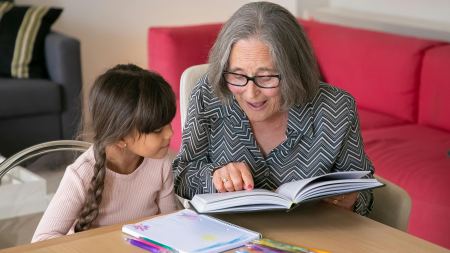 Ältere Frau sitzt mit Kind an Tisch und liest ein Buch. Auf dem Tisch liegen Heft und Stifte.