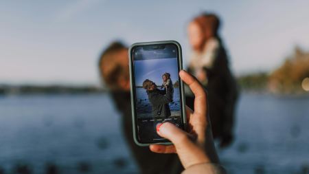 Eine Hand hält ein Handy fest, dieses ist auf einen Mann mit Kleinkind gerichtet.