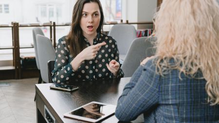 Zwei Frauen sitzen sich gegenüber an einem Tisch. Eine der beiden hat den Mund geöffnet und hebt die Finger vor sich. 