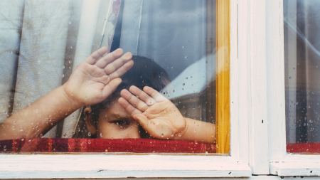 Kind hinter verschlossenem Fenster verschränkt die Arme vor dem Gesicht 