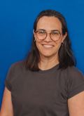 Portrait von Nadine Brexeler: Eine Frau mit schulterlangen braunen Haaren, Brille und braunem TShirt steht vor einem blauen Hintergrund und lächelt in die Kamera
